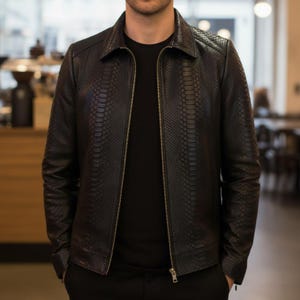 May include: A black leather jacket with a snakeskin texture. The jacket has a collared neckline, a zipper closure, and two front pockets. The jacket is worn over a black t-shirt.
