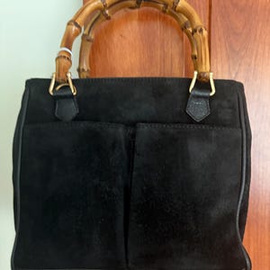 May include: A black suede handbag featuring two exterior front pockets and a curved bamboo handle. The handle is attached to the bag with gold-toned metal hardware. The bag has a structured rectangular shape with dark leather trim along the edges.