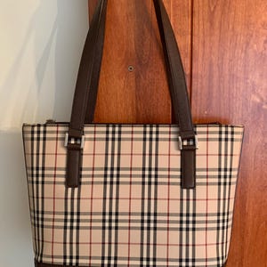 May include: A beige tote bag with a classic plaid pattern in black, red, and white. The bag has brown leather handles and trim, with silver-tone hardware. The base of the bag is a solid brown colour.