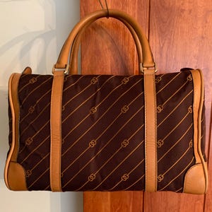 May include: Brown handbag with a repeating gold chain pattern and tan leather accents. The bag has two top handles and a rectangular shape. The bag's sides and base are trimmed with tan leather.