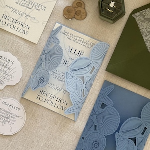 Pode incluir: Um conjunto de convites de casamento com cartolina azul-clara cortada a laser com padr&otilde;es intrincados de conchas e estrelas-do-mar. O conjunto inclui um cart&atilde;o de rece&ccedil;&atilde;o, um cart&atilde;o de detalhes, um cart&atilde;o de c&oacute;digo de vestimenta, um envelope verde-escuro e uma caixa de an&eacute;is com dois an&eacute;is dourados.