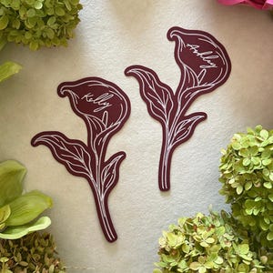 Peut inclure: Deux autocollants en forme de lys calla bordeaux profond avec des contours en ligne blanche. Chaque autocollant affiche un prénom en écriture cursive blanche, l'un indiquant Kelly et l'autre Ashley. Les autocollants sont plats et mats.