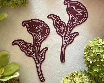 Op de afbeelding: Twee diepbordeauxrode calla lelie stickers met witte lijntekeningen. Elke sticker toont een naam in wit cursief schrift, waarbij de ene Kelly leest en de andere Ashley. De stickers zijn plat en hebben een gladde, matte afwerking.