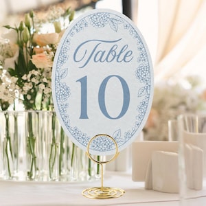 May include: An oval table number card featuring the text Table 10 in blue script and serif font, surrounded by a blue floral border. The card is held upright by a gold-colored metal wire stand with a circular base and a spiral top holder.