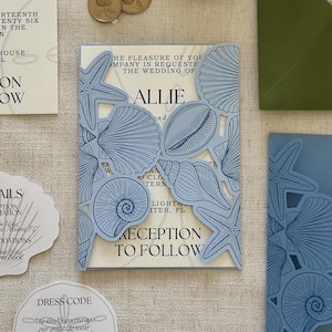 May include: A wedding invitation suite featuring a light blue laser-cut gatefold cover with intricate seashell and starfish illustrations. The cream-colored inner card displays black text including the name Allie and reception details.