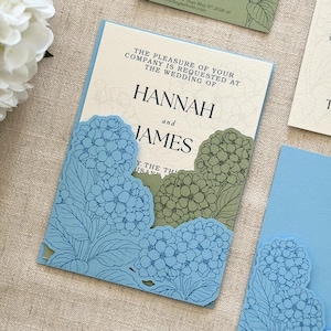 Puede incluir: Invitación de boda con un diseño floral azul y los nombres "Hannah y James". La invitación tiene un diseño recortado floral azul claro y un fondo color crema. También se ve papelería adicional a juego.