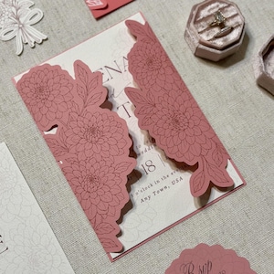 May include: Dusty rose wedding invitation with floral cutouts. The invitation features a laser-cut floral design and includes the date and time of the event. A ring box and other stationery items are also visible.