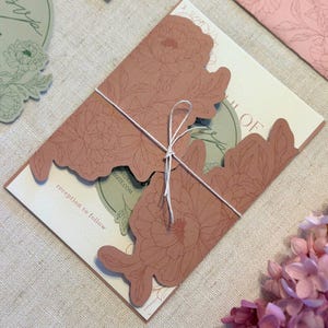 Könnte beinhalten: Eine Hochzeitseinladungssuite mit einem lasergeschnittenen floralen Design in Staubrosa. Die Einladung ist mit weißem Bindfaden gebunden und enthält den Text "reception to follow" und eine Website-Adresse. Zusätzliche Karten mit floralen Designs sind sichtbar.