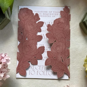 May include: A wedding invitation card featuring two die-cut, dusty rose-colored paper panels with intricate line-art rose and leaf patterns. The white cardstock underneath displays printed text including the words, The pleasure of your company is requested at the wedding of, and Reception to follow.