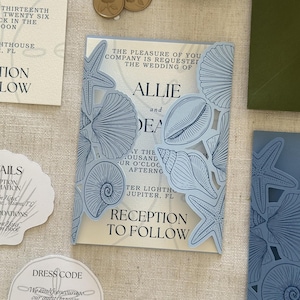 Pode incluir: Um conjunto de convites de casamento com uma capa dobr&aacute;vel azul-clara cortada a laser, decorada com ilustra&ccedil;&otilde;es de conchas e estrelas-do-mar. O cart&atilde;o interno exibe texto em preto com os nomes Allie e Dean, al&eacute;m de outros itens de papelaria a condizer.
