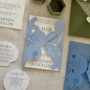 Pode incluir: Um conjunto de convites de casamento com capas de cartolina azul-claro cortadas a laser, com padr&otilde;es detalhados de conchas e estrelas-do-mar. Os encartes em tom creme exibem texto preto com o nome Allie e detalhes do evento.