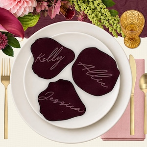 May include: Three dark burgundy, organic-shaped place cards featuring the names Kelly, Allie, and Jessica written in a light-coloured script font. These custom name tags are arranged on a white dinner plate, accompanied by gold-toned cutlery.