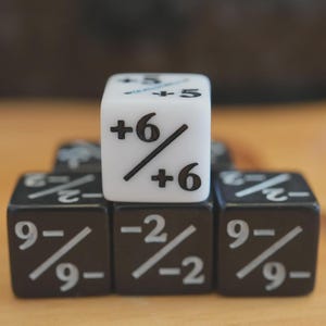 Peut inclure: Gros plan sur un ensemble de dés. Un dé blanc est empilé sur trois dés noirs. Le dé blanc affiche +6 et -5. Les dés noirs affichent 9- et -2. Les dés sont pour un jeu de société.