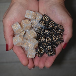 May include: A collection of dice in two colors, held in cupped hands. One group is white with gold markings, the other is black with gold markings. The dice are six-sided and feature symbols and numbers.