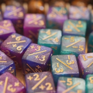 Könnte beinhalten: Eine Sammlung farbenfroher Würfel mit goldfarbenen Zahlen und Symbolen. Die Würfel sind eine Mischung aus Blau, Lila und Türkis, mit einem glitzernden, galaxieartigen Effekt. Die Würfel werden für Rollenspiele verwendet.