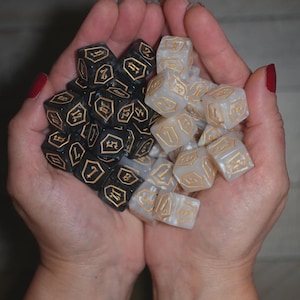May include: A handful of black and white polyhedral dice with gold-colored numbering. The dice are held in open hands, showcasing the contrasting colors and intricate details of the gaming accessories.