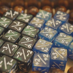 Könnte beinhalten: Eine Sammlung von Würfeln, einige in Grün und Schwarz, andere in Blau. Jeder Würfel hat weiße Markierungen, einschließlich Zahlen und Symbole, für Tischspiele. Die Würfel sind in einem Raster angeordnet.