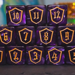 May include: A collection of purple and gold dice, each displaying a number from 2 to 12 within a shield design. The dice have a marbled effect, and the numbers are in a classic font. These dice are likely for tabletop games.