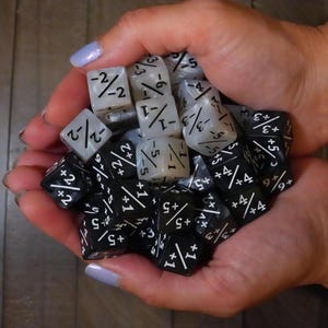 Peut inclure: Une poignée de dés, certains noirs et d'autres blancs translucides, sont tenus dans des mains en coupe. Les dés ont des marquages blancs, y compris des fractions et des signes plus. Dés pour jeux de société.