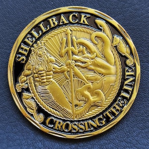 Può includere: Una moneta sfida circolare color oro e nero con un rilievo di Re Nettuno che tiene un tridente, circondato da creature marine. Il testo SHELLBACK e CROSSING THE LINE è impresso in lettere dorate sul bordo nero esterno.