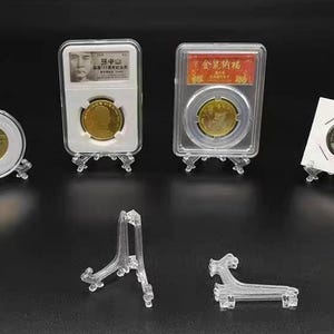 May include: Four collectible coins displayed in protective holders on small clear plastic stands. Two coins are in rectangular slabs, one in a round capsule, and one in a square cardboard holder. Two additional empty clear plastic display stands.