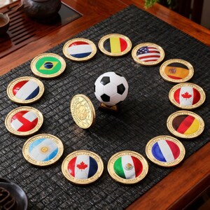 May include: A collection of circular gold-toned metal coins featuring various national flags and the text "FOOTBALL FEAST 2026" arranged around a central miniature black and white soccer ball. Each coin displays a distinct flag design.