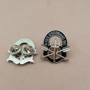 May include: A silver-coloured metal lapel pin featuring a central dagger crossed with two arrows, enclosed by a black arched border with the text DE OPPRESSO LIBER. The reverse side shows two metal pin back fasteners with circular clutch backs.