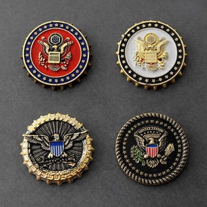 May include: Four circular metal pins featuring the Great Seal of the United States. Two pins have red and white backgrounds with gold eagles, while the other two feature black backgrounds with detailed gold and coloured eagle emblems and star borders.