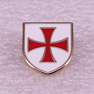 Puede incluir: Un pin de esmalte con forma de escudo que presenta un fondo blanco con una cruz roja llamativa centrada en la superficie. El pin está delineado en un tono dorado metálico, creando un borde distintivo que enmarca claramente el diseño.
