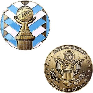 Puede incluir: Dos insignias redondas de metal. Una presenta un globo terráqueo con "Afganistán", sostenido por manos, sobre un fondo geométrico azul y blanco. La otra insignia tiene las palabras "American Embassy Kabul Afghanistan" alrededor de un águila.