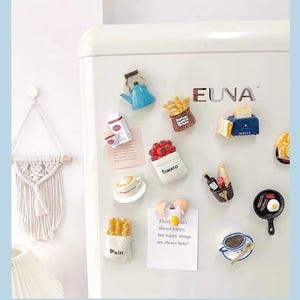 May include: A collection of colorful refrigerator magnets in various food shapes, including a blue teapot, a basket of fries, a tomato bag, a toaster, and a cup of coffee. A note says "Every isn't always happy, but happy things are always here!"