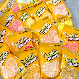 Pode incluir: Uma coleção de embalagens amarelas de Artkal Mini Beads. Cada embalagem contém contas em forma de coração em várias cores, incluindo branco, rosa, amarelo e laranja. As embalagens são rotuladas com o nome da marca e o número de contas.