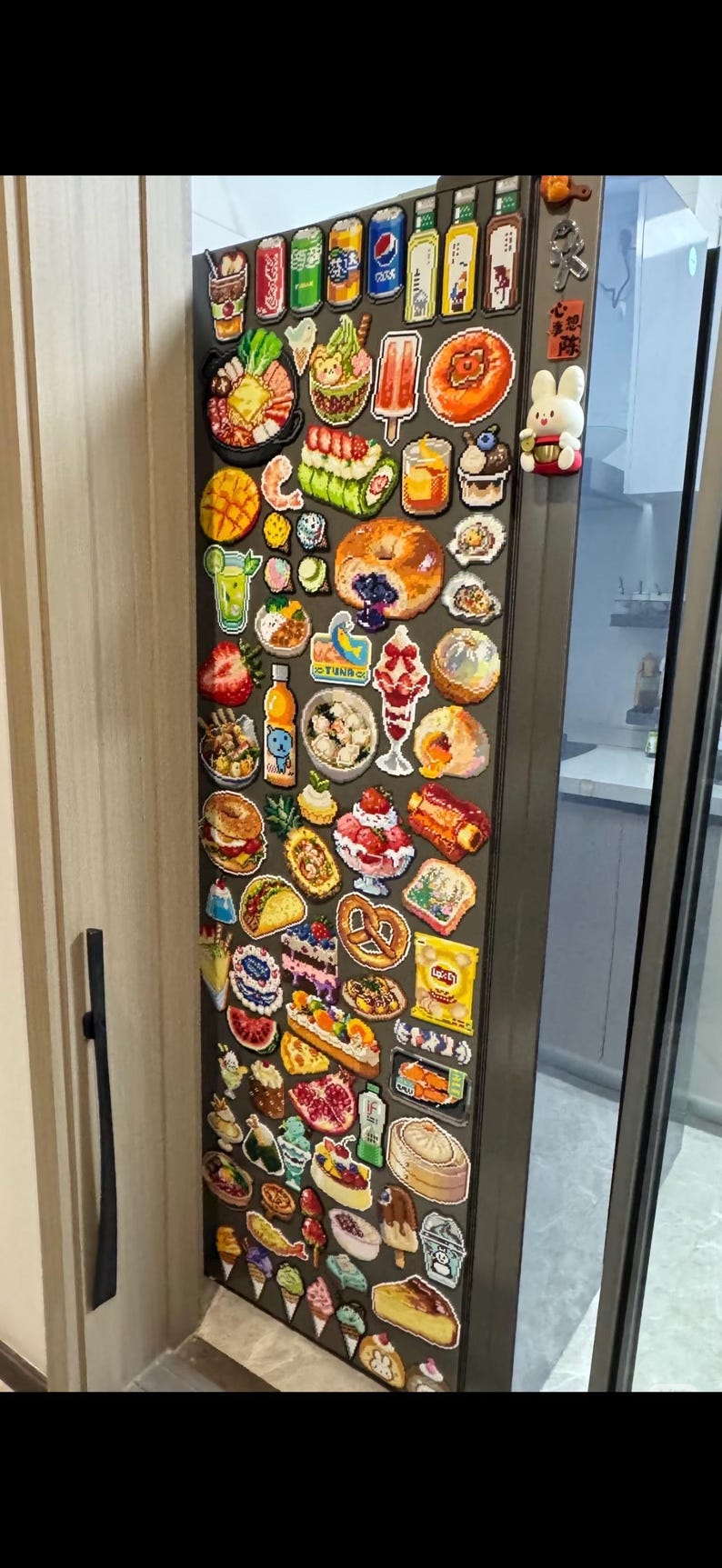 May include: A refrigerator door covered in colorful food-themed magnets. The magnets depict various foods and drinks, including soda cans, sushi, ice cream, and pastries. A cute bunny magnet is also visible.