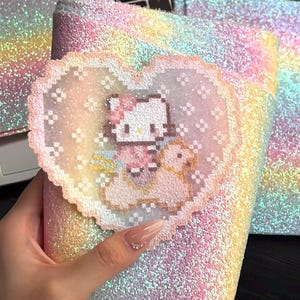 May include: A glittery notebook with a rainbow gradient design. The notebook features a heart-shaped embellishment with a pixelated Hello Kitty design. The cover and embellishment have a sparkly, iridescent finish, creating a whimsical aesthetic.