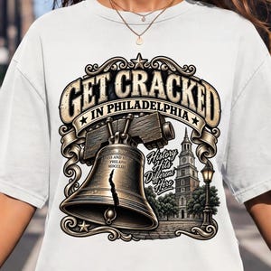 Könnte beinhalten: Ein weißes T-Shirt mit einem Grafikdruck der Liberty Bell mit einem deutlichen Riss, dem Text "GET CRACKED IN PHILADELPHIA" und "History Hits Different Here" sowie einer Illustration der Independence Hall und einer Straßenlaterne.