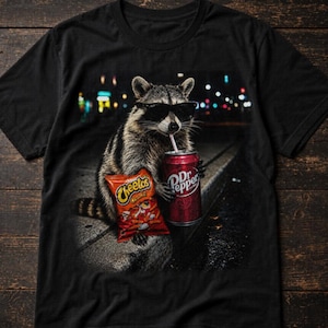 May include: A black t-shirt featuring a graphic print of a raccoon wearing black sunglasses. The raccoon is holding a red bag of Cheetos and a red can of Dr Pepper with a straw, set against a dark, out-of-focus background with city lights.