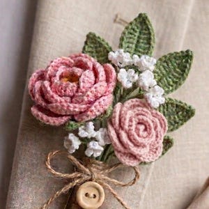 Op de afbeelding: Handgemaakte gehaakte corsage met twee roze rozen, witte vulbloemen en groene bladeren. Het boeket is vastgebonden met touw en een houten knoop. De tekst onderaan luidt "CROCHET WEDDING BOUTONNIERE PATTERN 4 FLOWER DESIGNS."