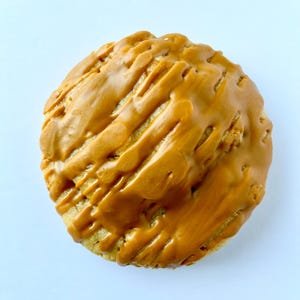 May include: A round cookie with a light brown base and a glossy, caramel-colored glaze drizzled across the top. The glaze has a textured, ridged appearance, creating a visually appealing treat. The cookie is set against a plain white background.