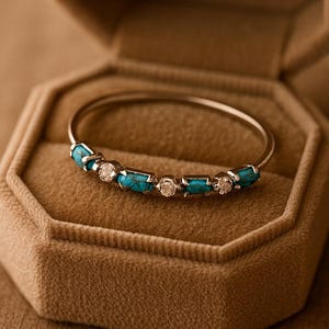 Op de afbeelding: Een zilveren armband met turquoise en diamant accenten ligt in een open, achthoekige, beige fluwelen sieradendoos. De armband heeft vijf turkooizen stenen en vier kleine diamanten.