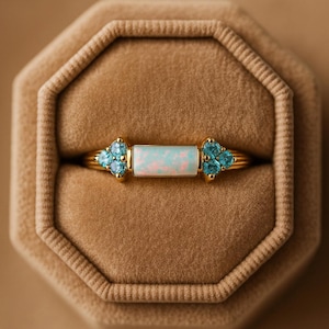 May include: A gold ring with a rectangular opal center stone and clusters of blue gemstones on each side. The ring is displayed inside a brown velvet ring box.