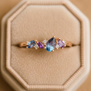 May include: A gold ring with a cluster of gemstones in blue, purple, and white. The ring features a central teardrop-shaped blue stone, surrounded by smaller stones. The ring is presented in a beige box.