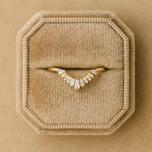 Könnte beinhalten: Ein goldfarbener Ring mit geschwungenem Design, besetzt mit mehreren kleinen, klaren, rechteckigen und dreieckigen Steinen. Der Ring wird in einer beigen Samtringbox präsentiert.