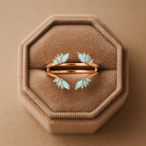 May include: A rose gold ring with a double band design. The ring features clusters of light blue opal-like stones arranged in a leaf-like pattern. The ring is displayed inside a brown velvet ring box.