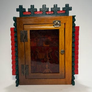 May include: A wooden cabinet with a hinged door and a decorative top featuring crosses. The cabinet has a dark wood frame, a glass door, and a red and green trim. The interior features a Day of the Dead theme.