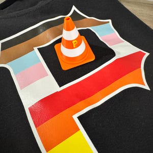 以下が含まれることがあります： 黒いTシャツ。白い縁取りのある大きな「P」の文字が虹色のグラデーションでデザインされています。オレンジと白のカラーコーンには黄色の「P」がプリントされており、Tシャツの上に置かれています。