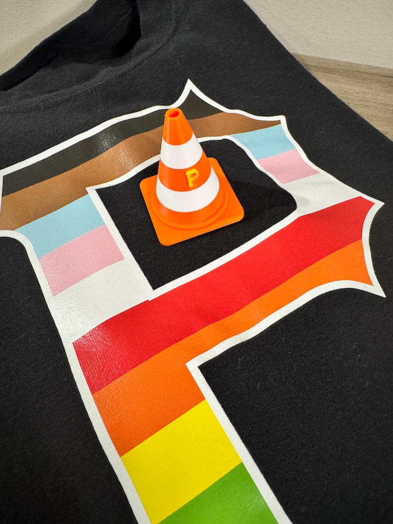 以下が含まれることがあります： 黒いTシャツ。白い縁取りのある大きな「P」の文字が虹色のグラデーションでデザインされています。オレンジと白のカラーコーンには黄色の「P」がプリントされており、Tシャツの上に置かれています。