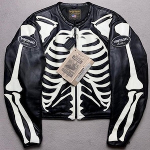 May include: Black leather jacket with a white skeleton design. The jacket has a zip closure, a round collar, and the "Vanson Leathers" logo on the sleeves. A tag with text hangs from the zip.