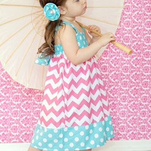 May include: A young girl in a colorful dress with a chevron pattern and polka dot accents. She is holding a cream-colored parasol and has a blue flower hair clip. The dress is pink, white, and turquoise.