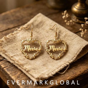 May include: A pair of gold-toned drop earrings featuring a heart-shaped frame made of barbed wire. The name Maria is written in a cursive script font across the center of each heart. The earrings are suspended from thin gold-toned metal hooks.