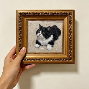 May include: An oil painting of a black and white cat with yellow eyes, depicted in a textured impasto style. The cat is shown in a reclining position against a neutral background. The artwork is encased in an ornate, metallic gold frame.